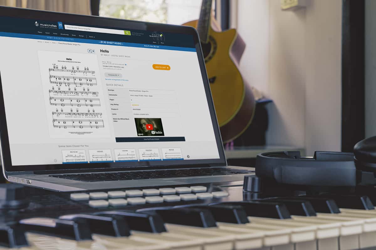 A laptop displaying digital sheet music sits on top of a synthesizer with a guitar in the background
